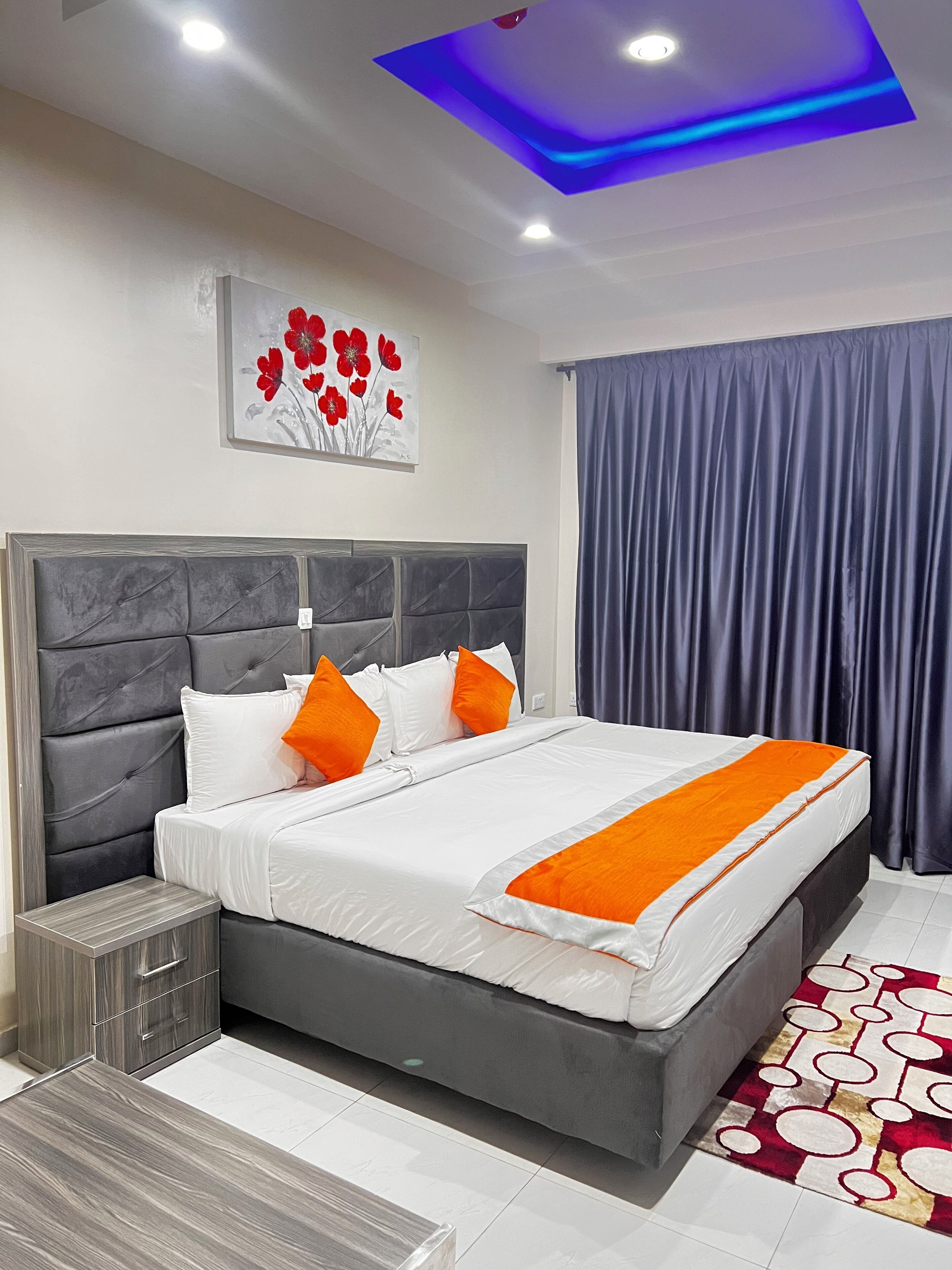 A modern hotel room with a large bed, grey headboard, white bedding, orange pillows, a grey nightstand, a small table, a red and white patterned rug, a blue ceiling light, and a framed artwork of red flowers on the wall.
