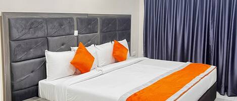 A modern hotel room with a large bed, grey headboard, white bedding, orange pillows, a grey nightstand, a small table, a red and white patterned rug, a blue ceiling light, and a framed artwork of red flowers on the wall.