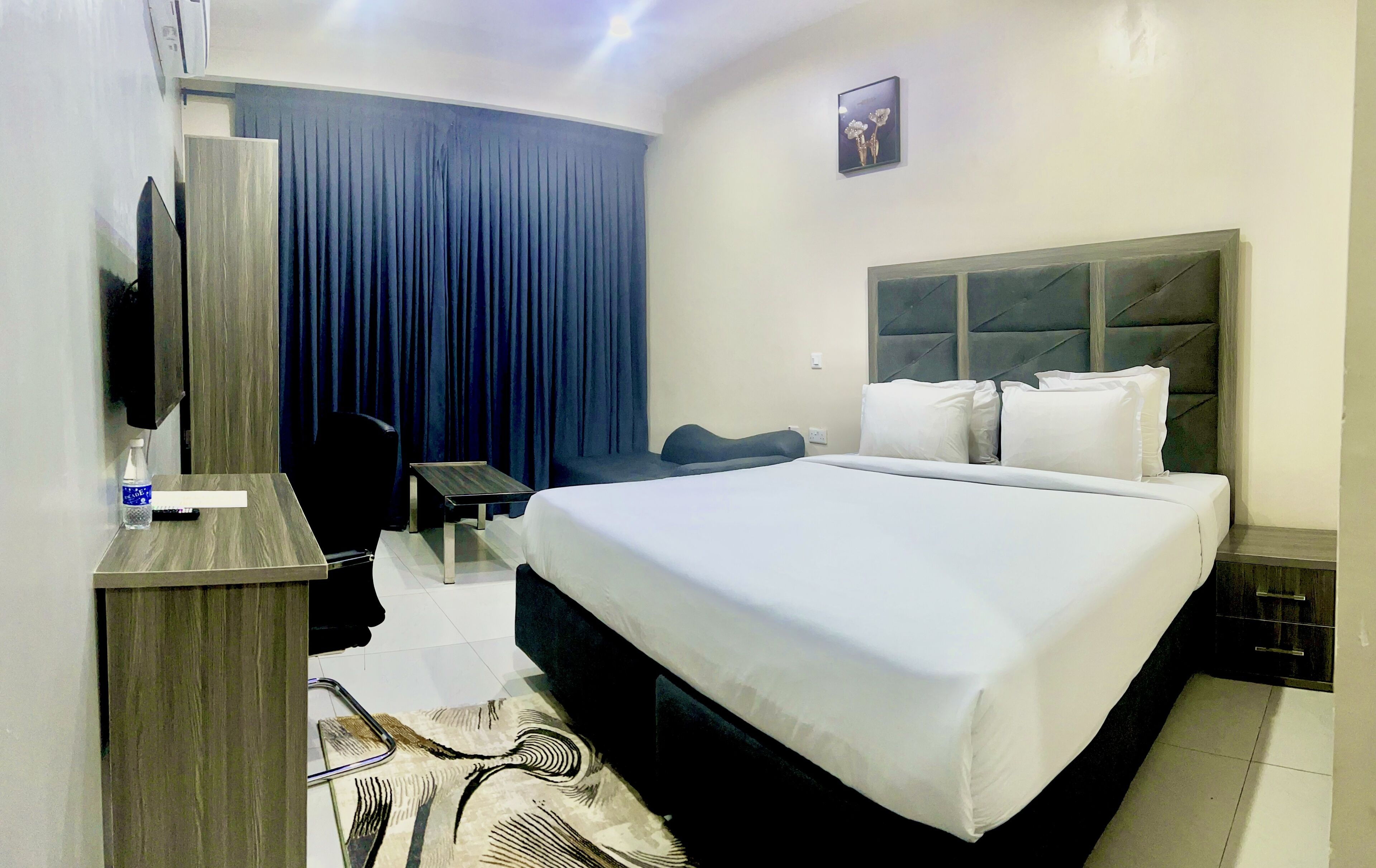 A hotel room with a large bed, a flat-screen TV mounted on the wall, a desk with a chair, a water bottle, and a remote control.