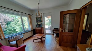 Ein geräumiges Zimmer mit Holzboden, einem roten Sessel, einem Holztisch und einem großen Fenster mit Blick auf Grünflächen.