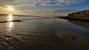 Ein ruhiger Strand mit blauem Himmel, einem sichtbaren Kondensstreifen und einer Windkraftanlage in der Ferne.