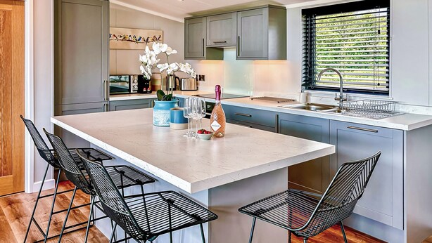 A modern kitchen with a central island, bar stools, and a window with blinds. - Royal Forest Pet Friendly Lodge (Coleford)