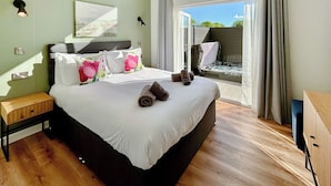 A hotel room with a large bed, a small bedside table, a nightstand, a painting on the wall, and a view of the outdoors. - Royal Forest Pet Friendly Lodge (Coleford)