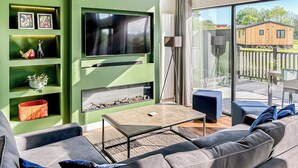 A modern living room with a green accent wall, a flat-screen TV, a sofa, a coffee table, and a view of a wooden house through large windows. - Royal Forest Pet Friendly Lodge (Coleford)