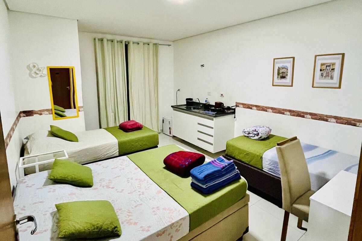 A hotel room with two beds, a desk, a chair, a small kitchenette, and a window with curtains.