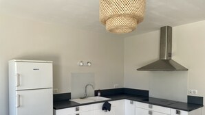 Una cocina moderna con armarios blancos, encimera negra, refrigerador blanco, campana extractora de acero inoxidable y lámpara de techo tejida.