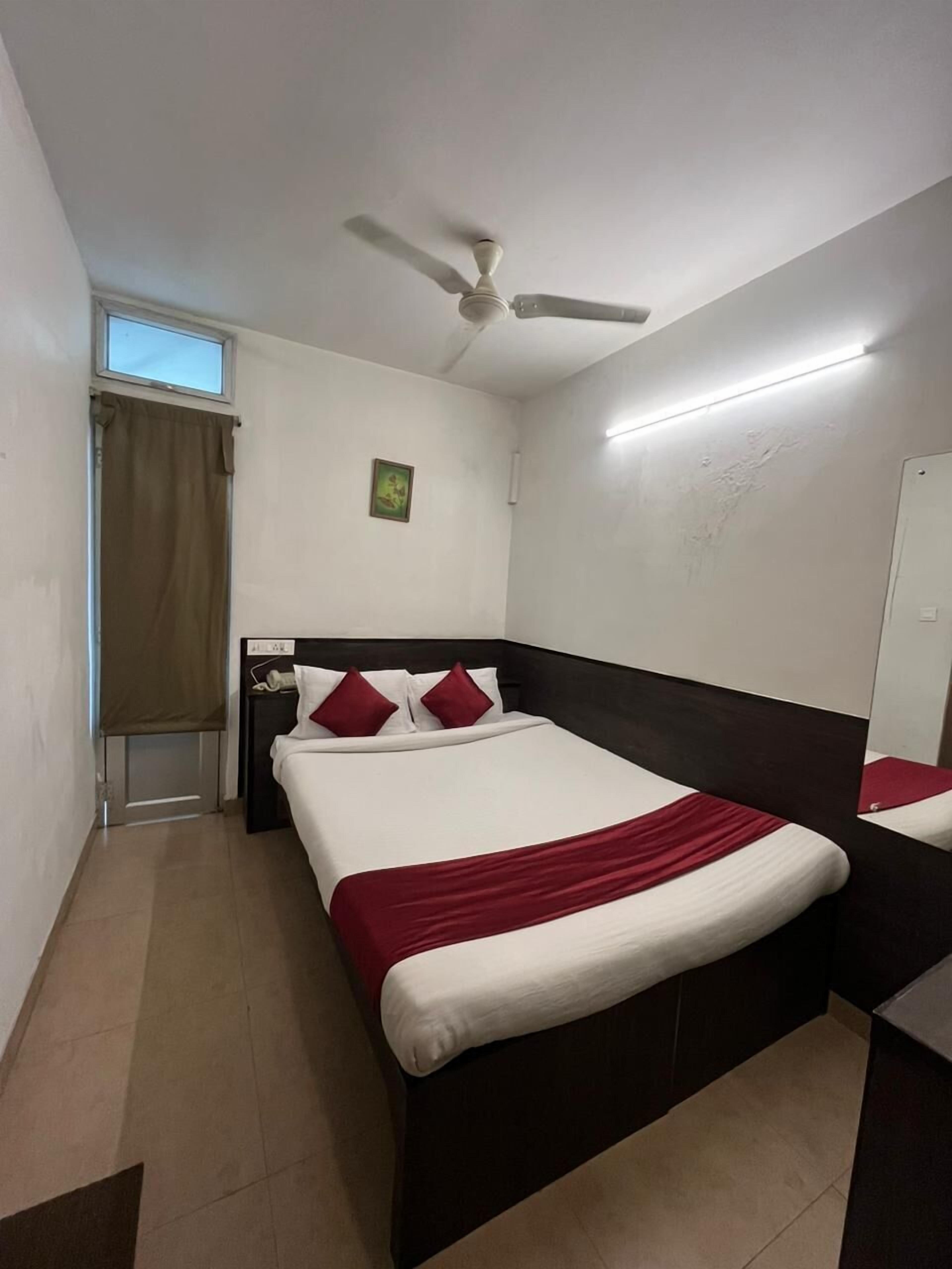 Habitación de hotel ordenada con cama, mesita de noche, ventilador de techo, ventana con cortinas y un pequeño cuadro en la pared.