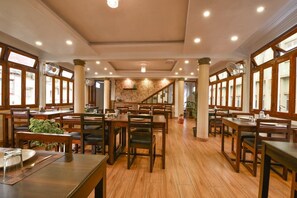 A spacious restaurant with wooden tables and chairs, large windows, and a staircase leading to an upper level. - Hotel Indiana (Shillong)