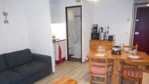 Een gezellig, compacte leefruimte met een bank, een houten zitbankje, een eettafel met stoelen, een keukenhoek met een magnetron, koffiezetapparaat en een kastje.