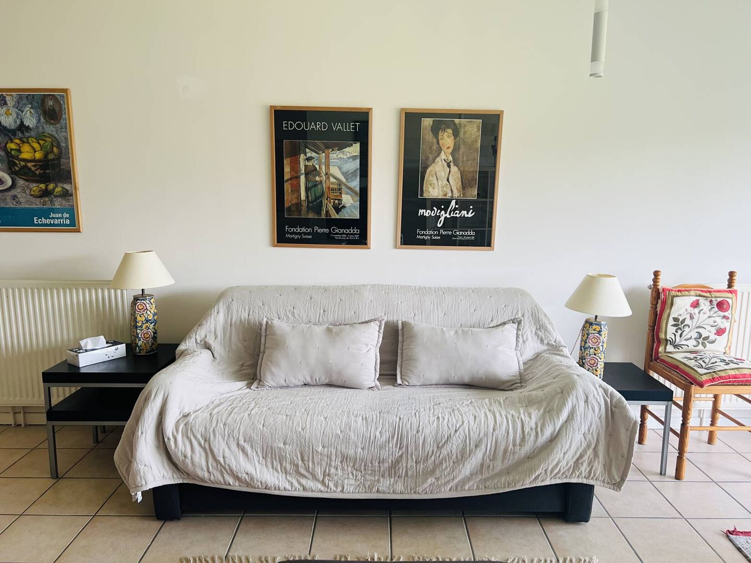 A room with a large bed, two bedside tables, and a chair. There are three framed artworks on the wall.