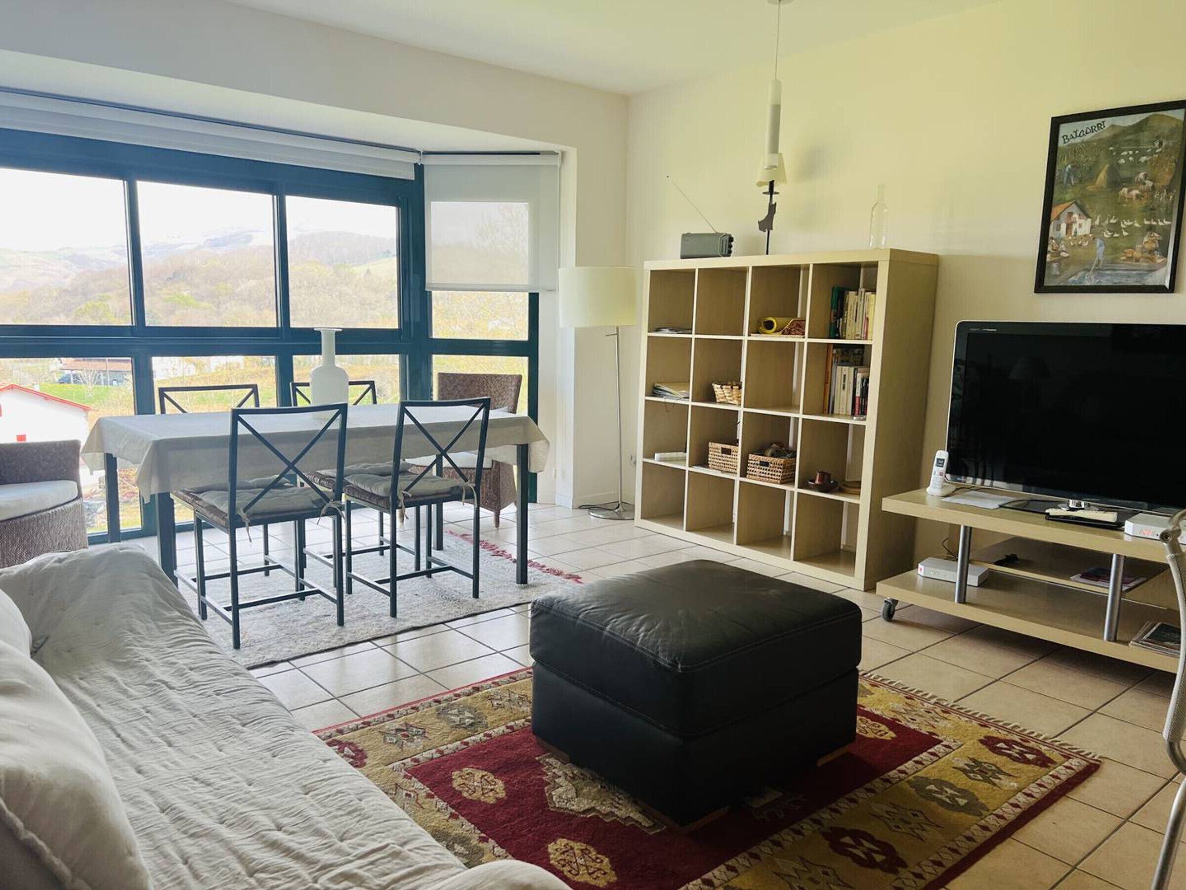 Een woonkamer met een bank, poef, eettafel, stoelen, een boekenkast, een televisie en een groot raam met uitzicht op de omgeving.