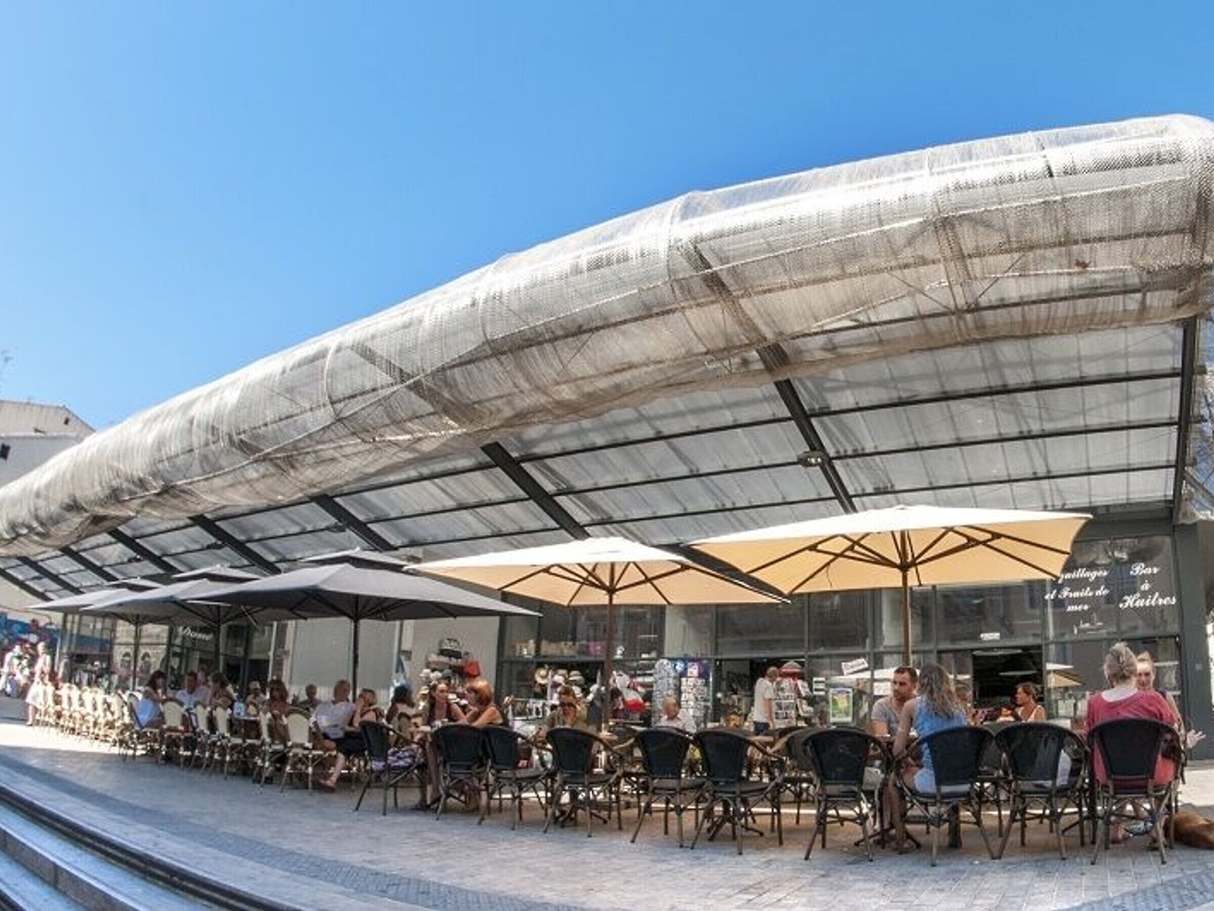 An outdoor café with tables and chairs under a large, transparent roof structure.