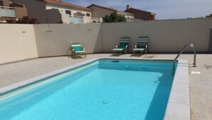Et rektangulært swimmingpool med krystalklart, blåt vand, omgivet af en lav, hvid mur. To liggestole med blå hynder og gyldne stel, og en ståltrappe til poolen på højre side.
