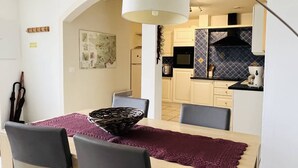 A dining area with a table set for four, a chandelier, and a view into a kitchen with modern appliances and a backsplash. - Charming T2 apartment in Cambo, city center, small terrace, Internet, pets allowed (Cambo-les-Bains)