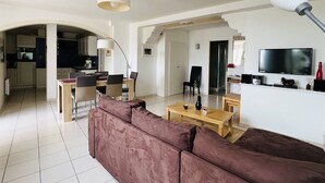A modern living room with a brown sofa, a wooden coffee table, and a TV on the wall. There is a kitchen visible through an archway. - Charming T2 apartment in Cambo, city center, small terrace, Internet, pets allowed (Cambo-les-Bains)