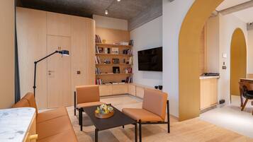 A modern living room with a sofa, a coffee table with a fruit bowl, a bookshelf, and a TV mounted on the wall.