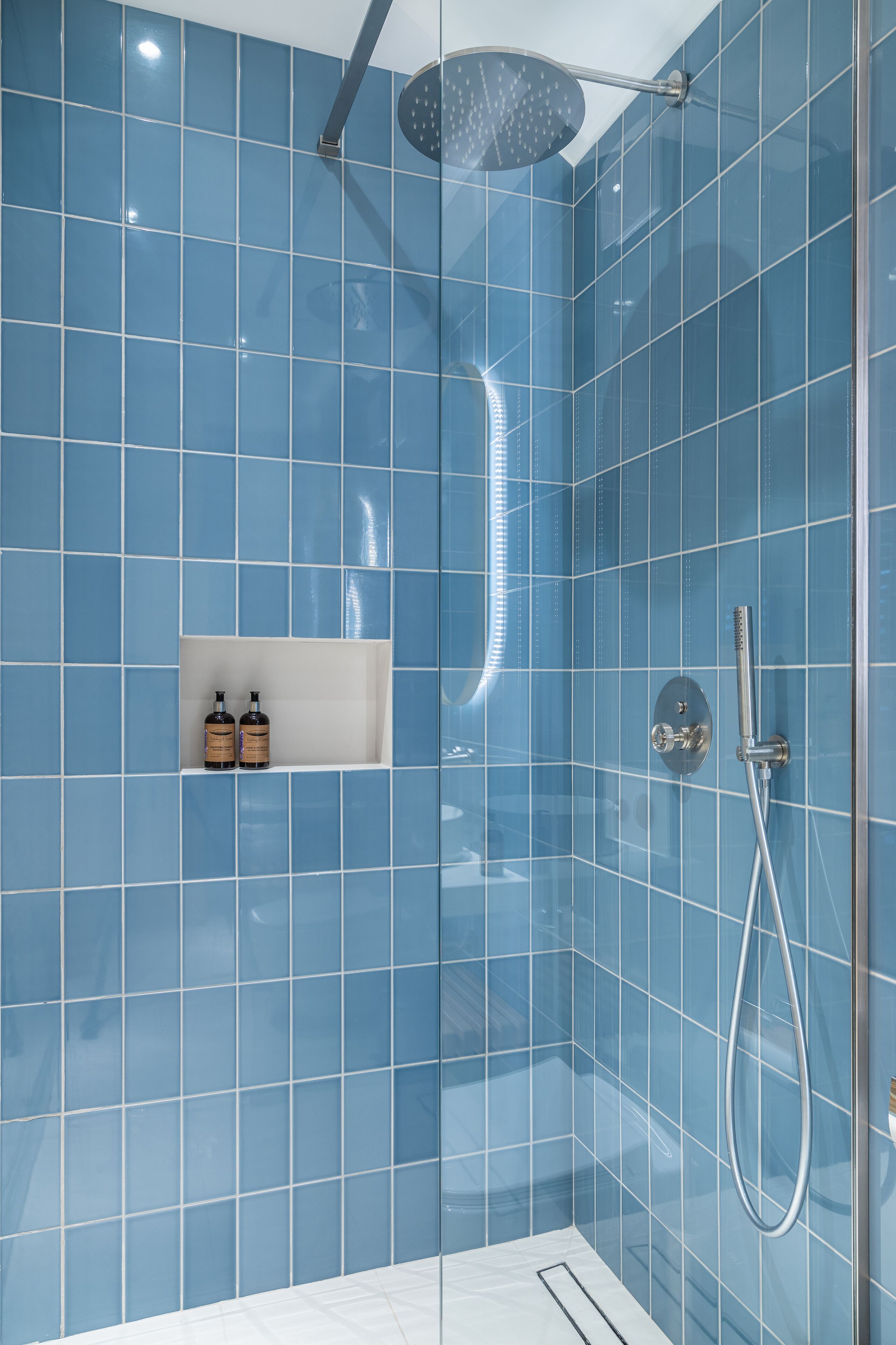 Un baño moderno con azulejos azules, cabezal de ducha y estante con artículos de aseo.
