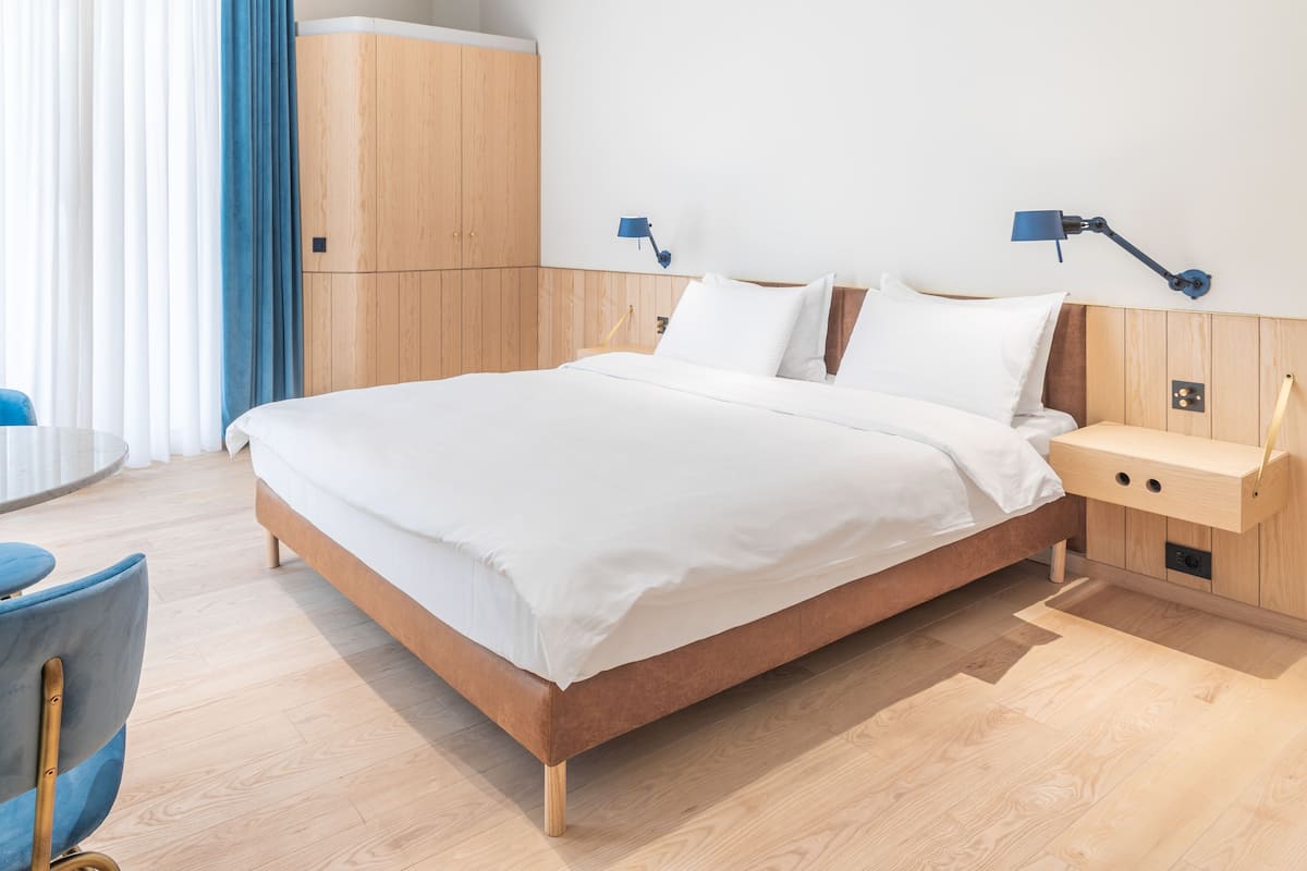 Une chambre d’hôtel moderne avec un lit en bois, deux tables de chevet, une armoire et une chaise bleue.