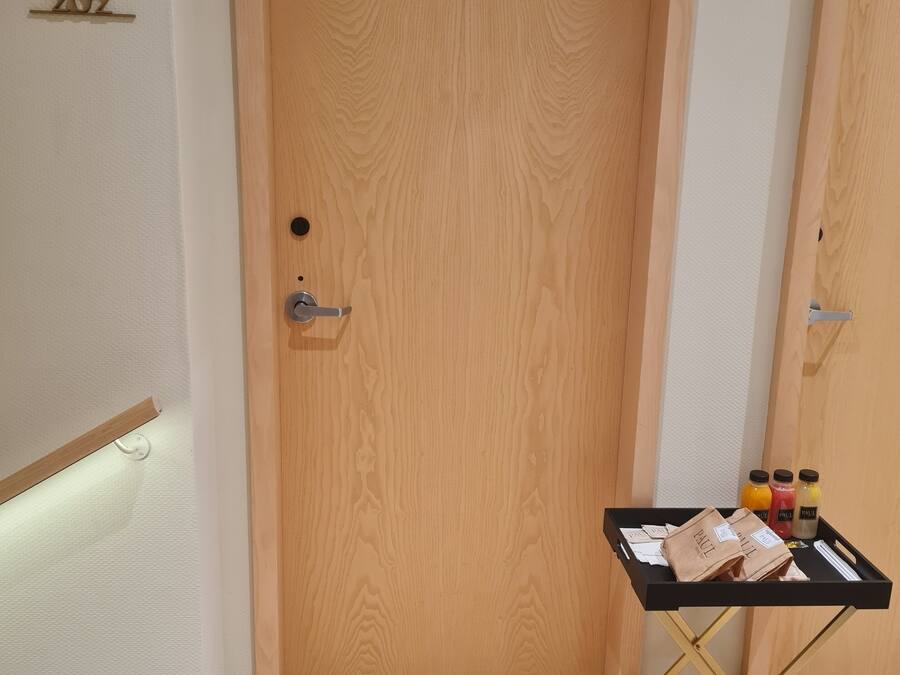 Una puerta de madera con una placa numérica, una mesa pequeña con bolsas y botellas, y un suelo alfombrado.