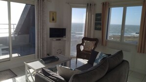 En stue med sofa, sofabord, fjernsyn og havudsigt gennem store vinduer.