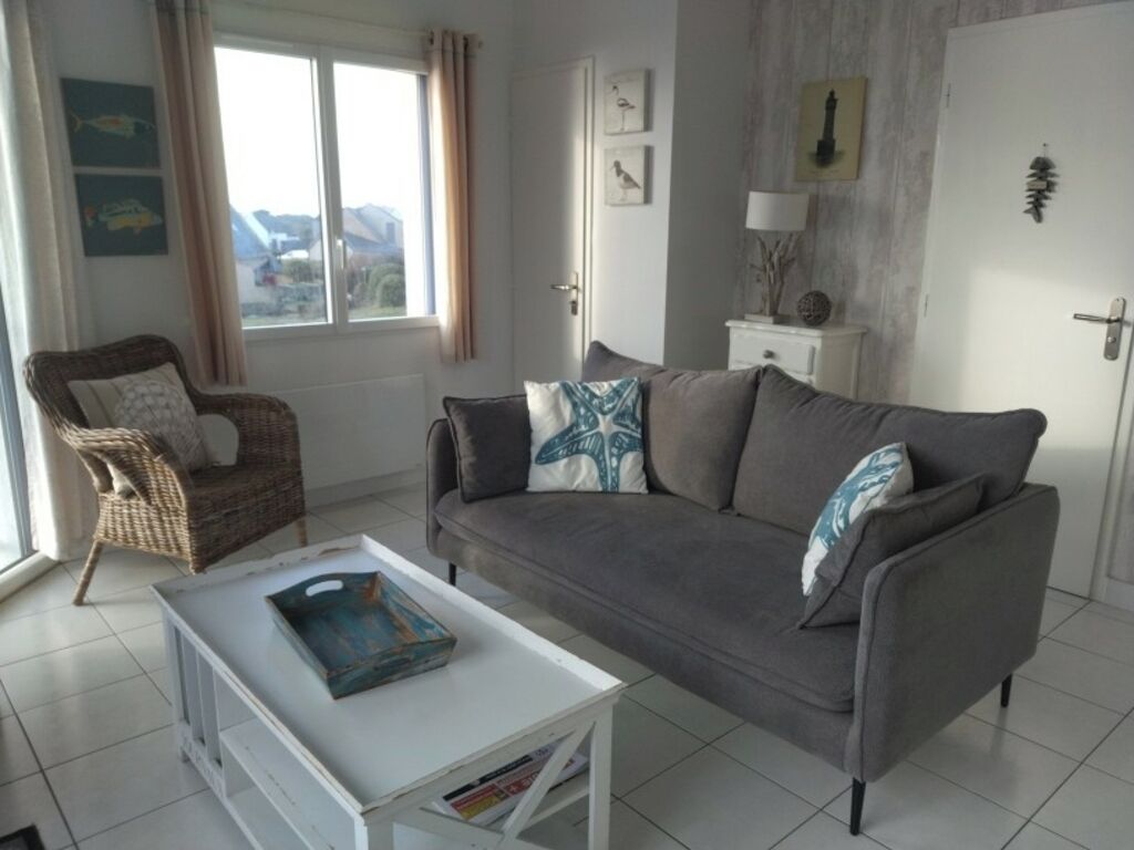 Een woonkamer met een grijze bank, een witte salontafel en een rieten stoel. Er is een raam met gordijnen, een deur en wanddecoratie.