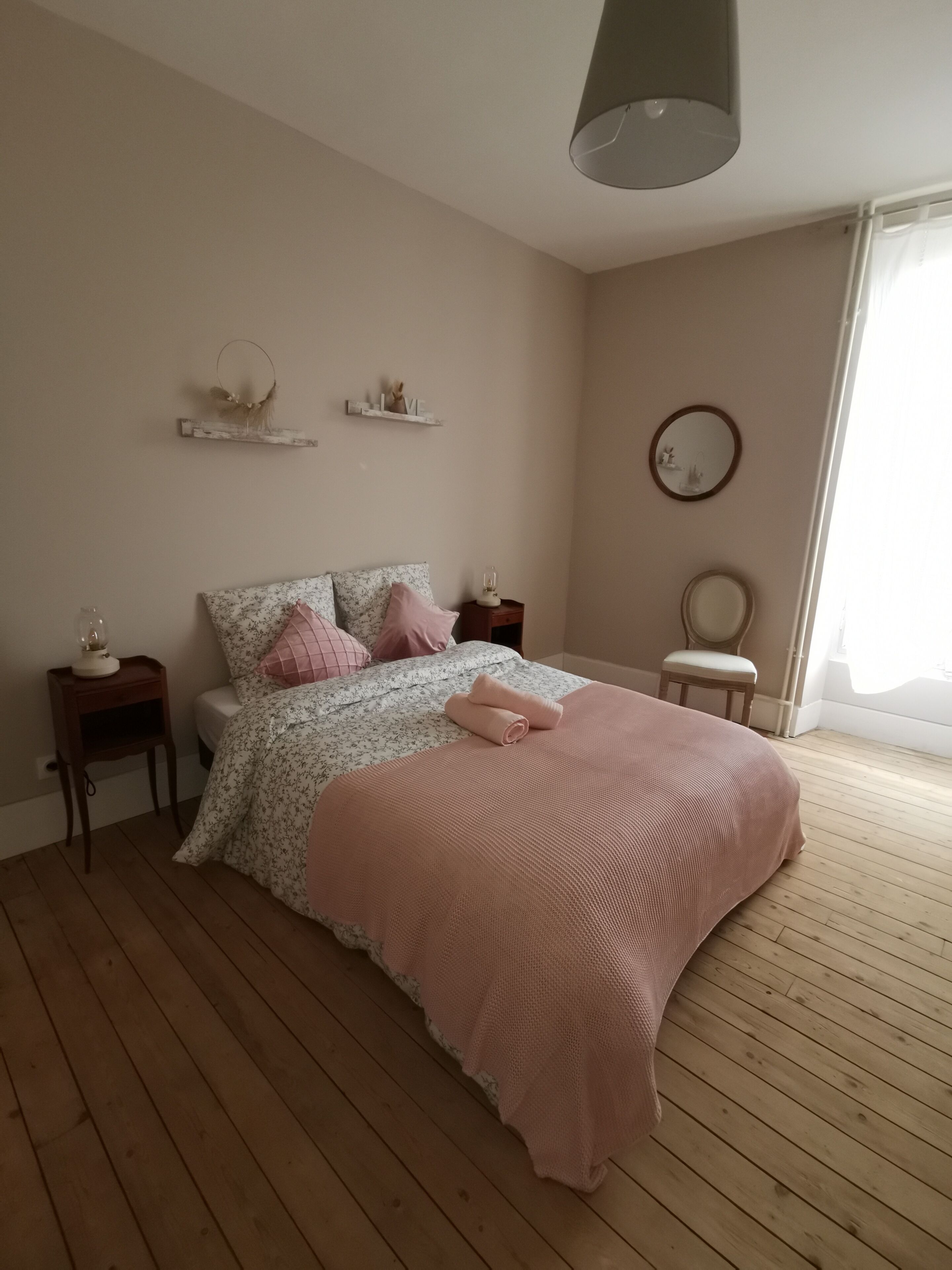 A bedroom with a wooden floor, a bed with floral bedding, a pink blanket, bedside tables with lamps, and a mirror on the wall.