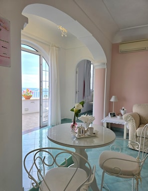 Una habitación con una mesa redonda, dos sillas, una mesita con tetera y taza, un florero con flores, y vistas al mar a través de grandes ventanales.