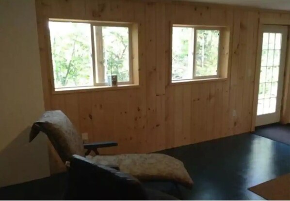 A room with wooden panel walls, two windows with views of greenery, a black chair with a cushion, and a dark floor. - Oceanfront lodge with all the amenities for a family vacation (Ellsworth)