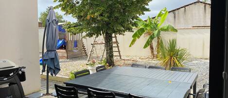 Un patio con mesa y sillas, una parrilla, un parque infantil y una palmera de plátano.