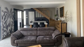 Una sala de estar moderna con un sofá gris, un comedor con mesa y sillas, una escalera y una cocina con armarios de madera y una planta en maceta.