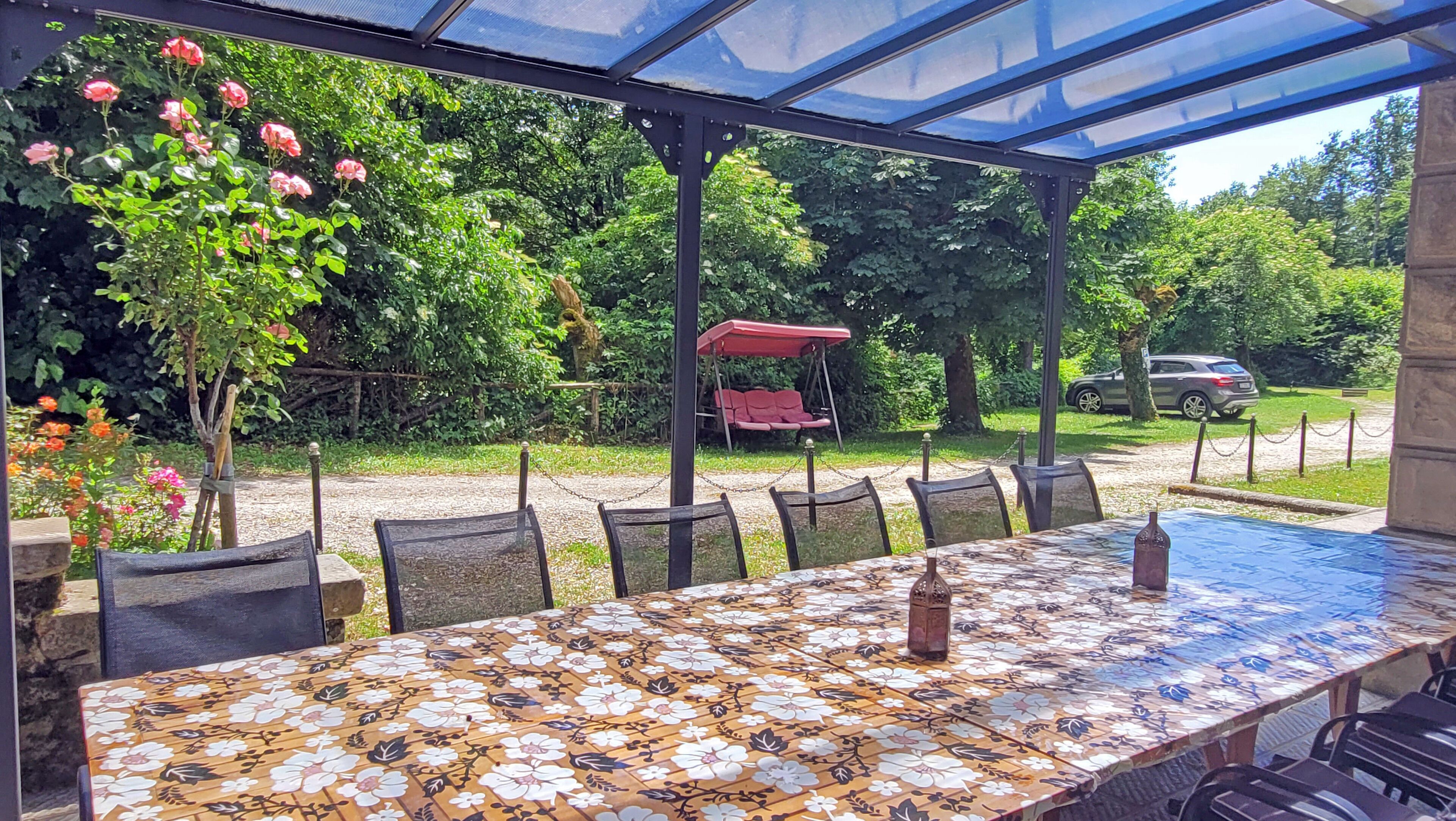 Un patio techado con una mesa, sillas, una hamaca y vistas a un coche y vegetación.