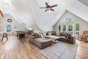 A spacious loft with a high ceiling, a pool table, a large sectional sofa, and a view of the outdoors through large windows.