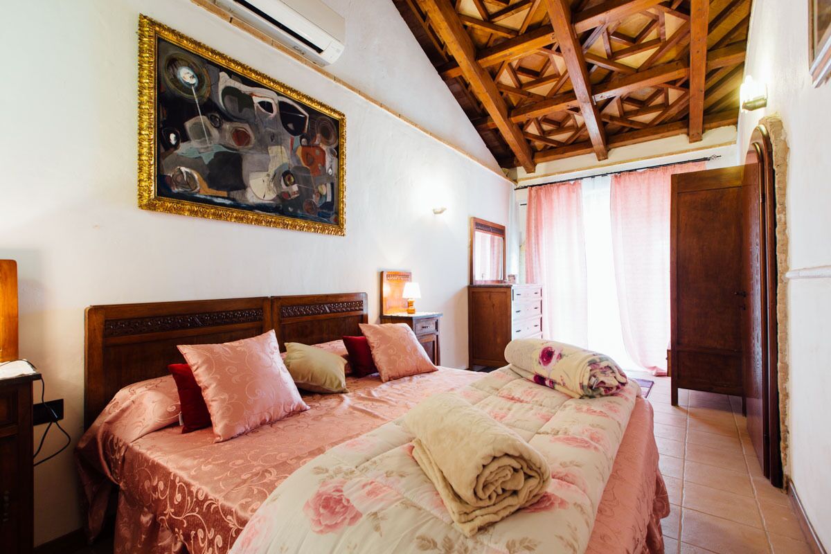 A bedroom with a wooden ceiling, a large bed with floral bedding, a painting on the wall, and a nightstand with a lamp.