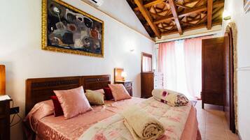 A bedroom with a wooden ceiling, a large bed with floral bedding, a painting on the wall, and a nightstand with a lamp.