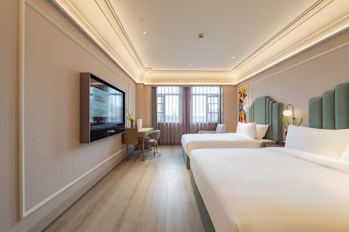 Une chambre d’hôtel équipée d’une télévision à écran plat, d’un bureau avec une chaise, de deux lits et d’une grande fenêtre avec des rideaux.