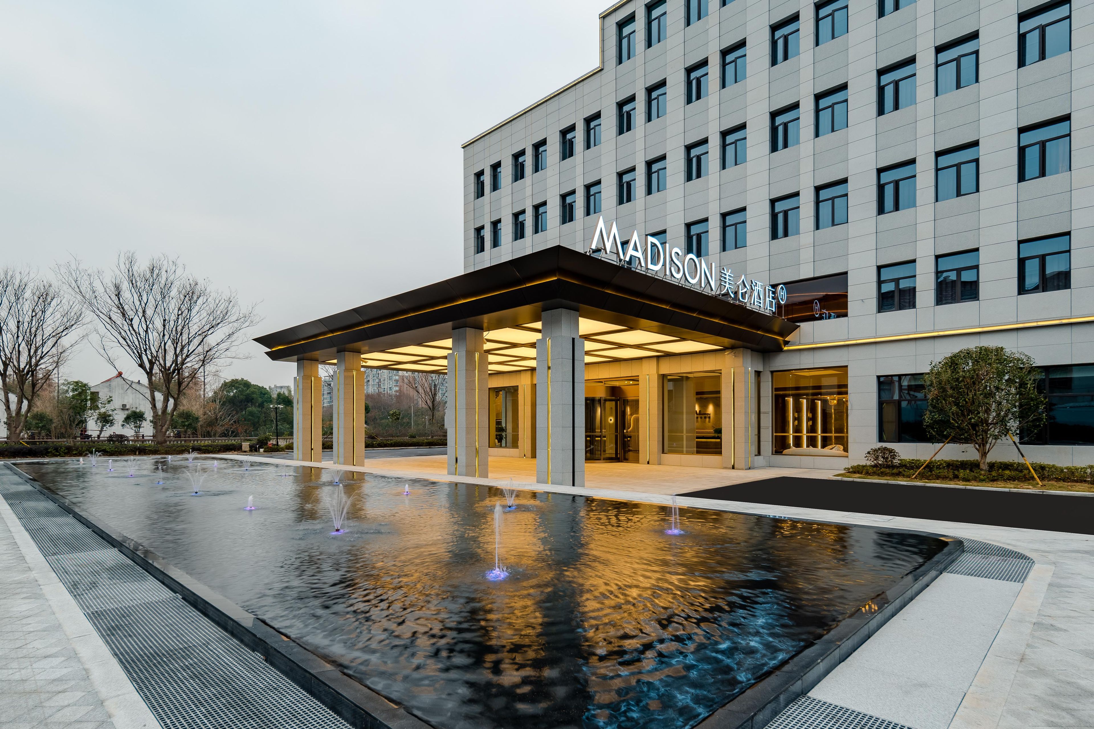 Entrada moderna de hotel con un área exterior techada, una fuente de agua y un edificio con múltiples ventanales.