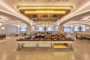 Un hall d’hôtel spacieux avec un buffet de petit-déjeuner central, des coins salon et de grandes fenêtres.