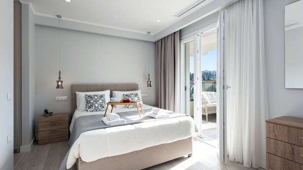 A modern bedroom with a large bed, a nightstand, a small table with towels, and a view through a sliding glass door. - PELAGIA SUITES (Corfu)