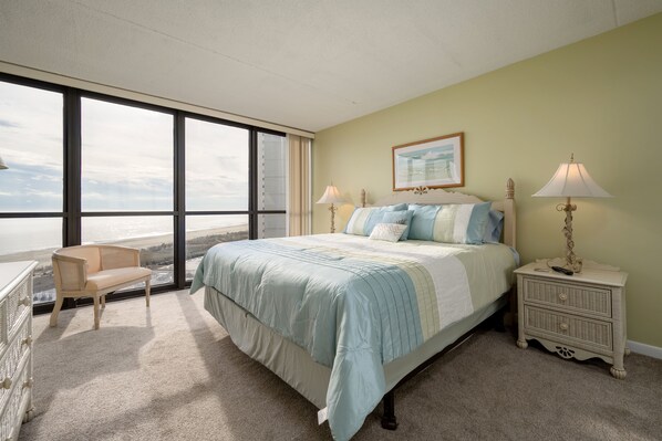 A bedroom with a large window, a bed with a light blue and white bedspread, a nightstand with lamps, and a chair.