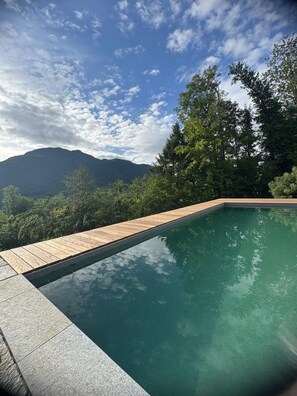 A swimming pool with a wooden deck, surrounded by lush greenery and a mountain in the background.