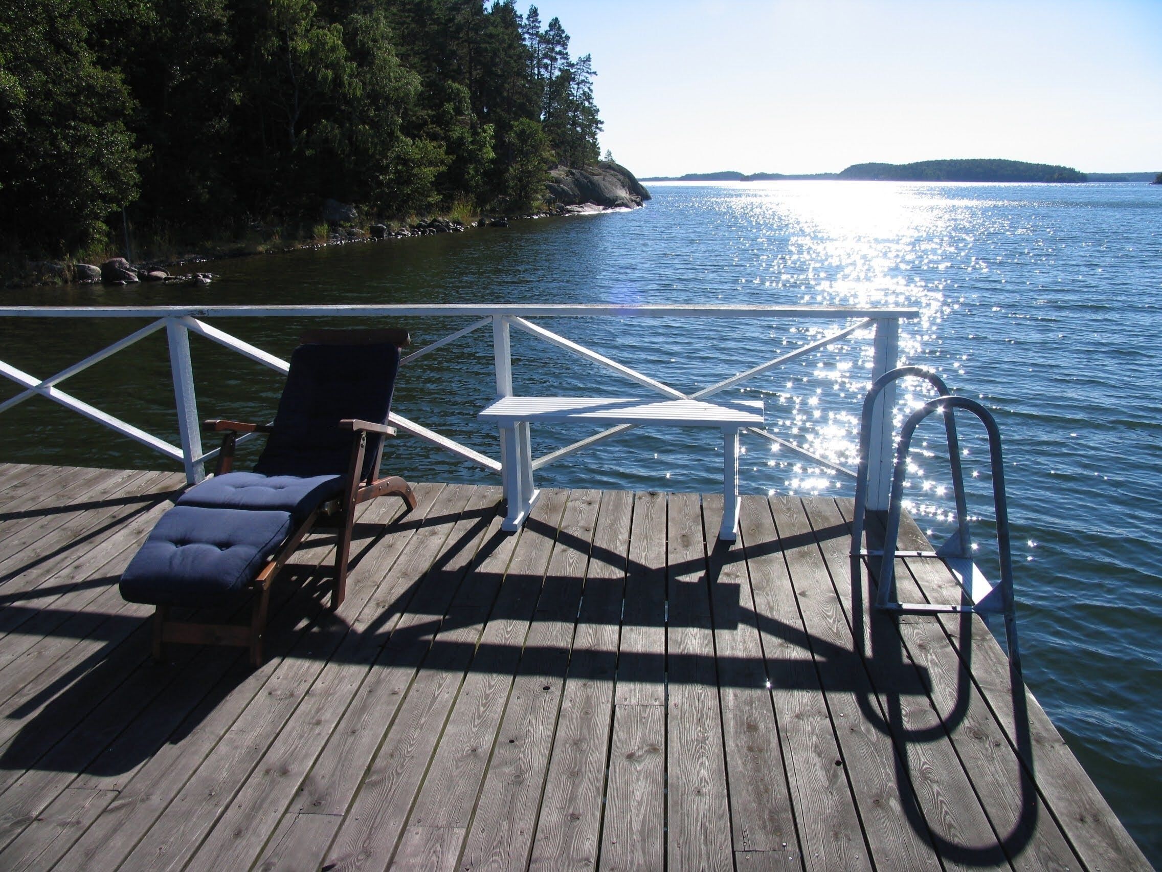 A wooden deck with a lounge chair and a table, overlooking a calm lake with a forested shoreline.