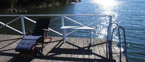 A wooden deck with a lounge chair and a table, overlooking a calm lake with a forested shoreline.