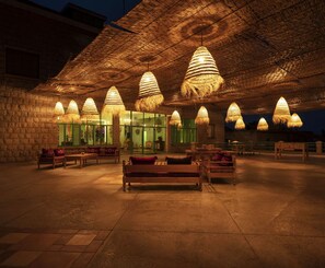 An outdoor seating area with thatched umbrellas and hanging lights, a stone wall, and a glass door leading inside. - Al Fundok (Maasser Al Chouf)