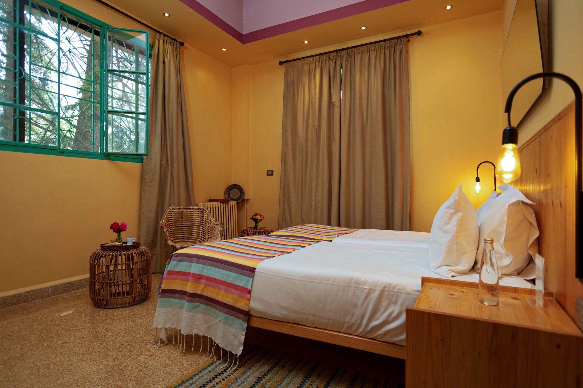 Um quarto com uma cama, uma mesa de cabeceira com abajur, uma cadeira de vime, uma pequena mesa com um vaso e uma janela com cortinas.
