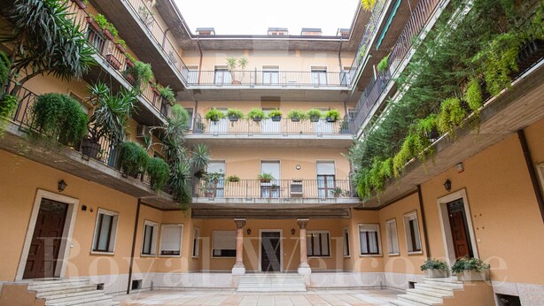 Un edificio de varios pisos con balcones decorados con plantas, una escalera central y un patio adoquinado.