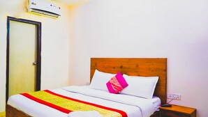 Une chambre d’hôtel avec une tête de lit en bois, un lit avec une literie blanche et une couverture rayée rouge et jaune, un oreiller rose et un climatiseur fixé au-dessus de la porte.