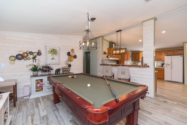 A pool table with a green felt surface, a wooden bar, and a hanging light fixture in a room with wooden flooring and a kitchen in the background.