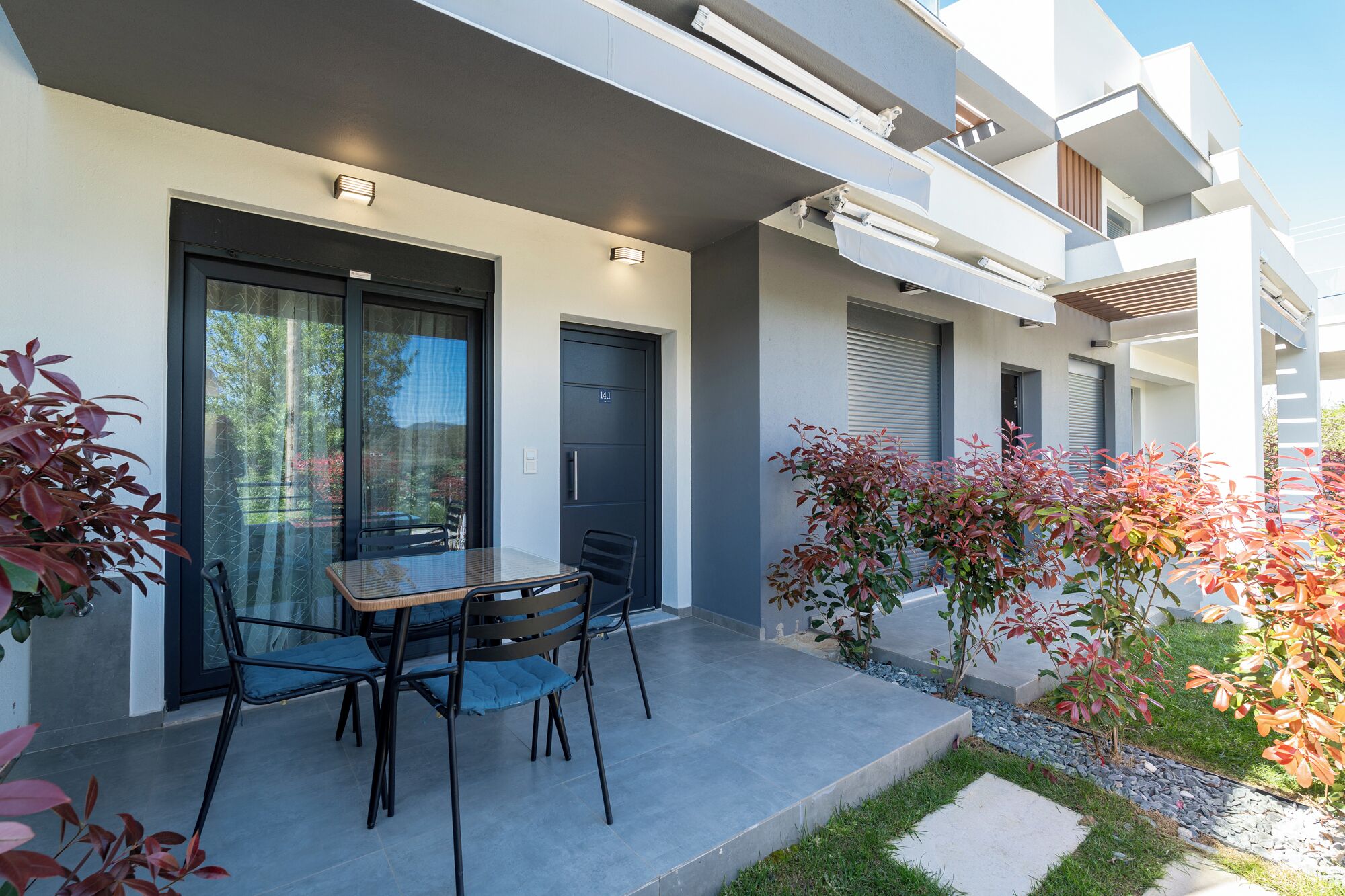A modern building with a glass door, a table with chairs, and a small garden area.