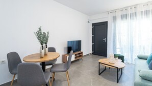 A modern living room with a round wooden dining table, grey upholstered chairs, a small side table with a vase, a television on a wooden stand, a dark door, and a green sofa. - Thalassa-One Bedroom Apartment (Kavala)