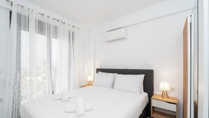 A hotel room with a bed, two bedside tables with lamps, a window with sheer curtains, and an air conditioning unit on the wall. - Thalassa-One Bedroom Apartment (Kavala)